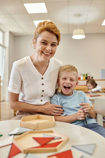 Веселый учитель обнимает мальчика рядом с деревянной игрой на столе в школе Монтессори на заднем плане — стоковое фото