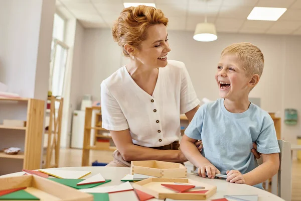 Carefree teacher hugging boy near wooden game on table in montessori class — Stock Photo