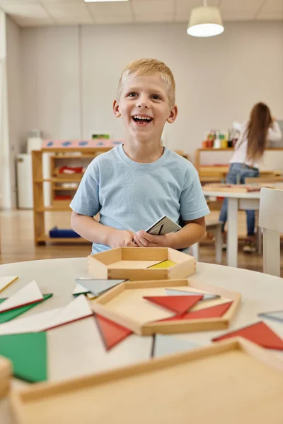 Niño alegre sosteniendo triángulo de madera y mirando hacia otro lado en clase en la escuela montessori - foto de stock