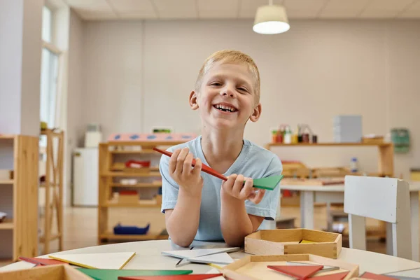 Веселый мальчик держа деревянную часть игры и глядя на камеру в классе в Монтессори школы — стоковое фото