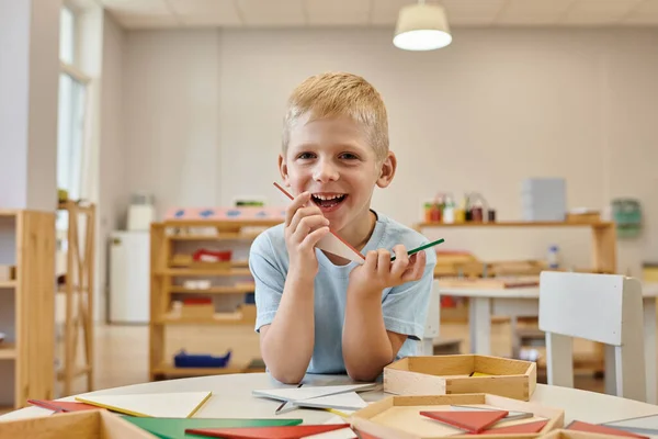 Menino alegre segurando triângulos durante o jogo em sala de aula na escola montessori — Fotografia de Stock