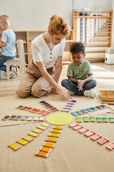 Professor falando com o menino afro-americano perto de tijolos de madeira coloridos no chão na escola montessori — Fotografia de Stock