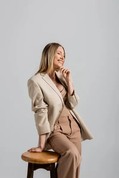 Allegra donna bionda in abito formale appoggiata sulla sedia e toccante mento su sfondo grigio, moda — Foto stock
