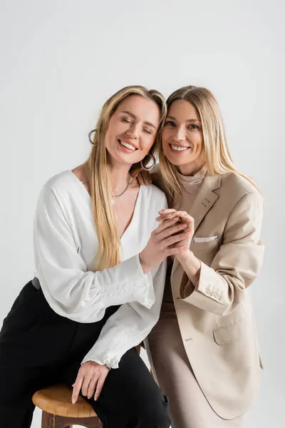 Two attractive elegant sisters sitting on chairs smiling and holding hands, fashion and style — Stock Photo