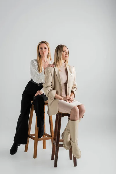 Atractivas hermanas elegantes en ropa formal sentadas en sillas, mano sobre hombro, moda y estilo - foto de stock
