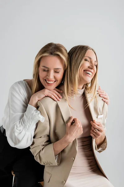 Beautiful stylish sisters in formal outfits beaming sitting on chairs and hugging, fashion concept — Stock Photo