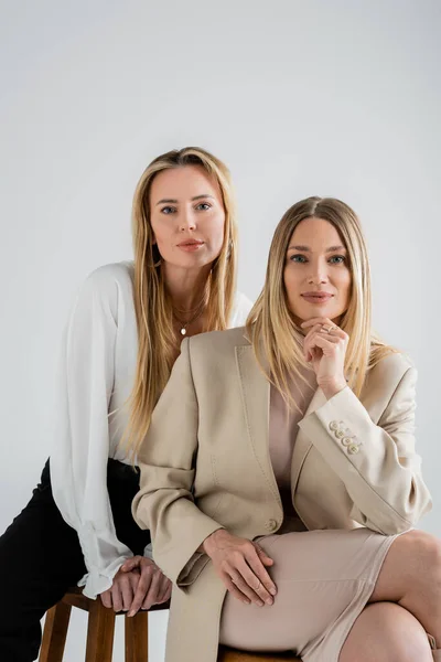Pretty and elegant sisters in formal wear sitting on chairs looking at camera, fashion concept — Stock Photo