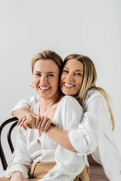 Retrato de irmãs loiras elegantes sentadas em cadeiras abraçando e sorrindo, estilo e moda — Fotografia de Stock