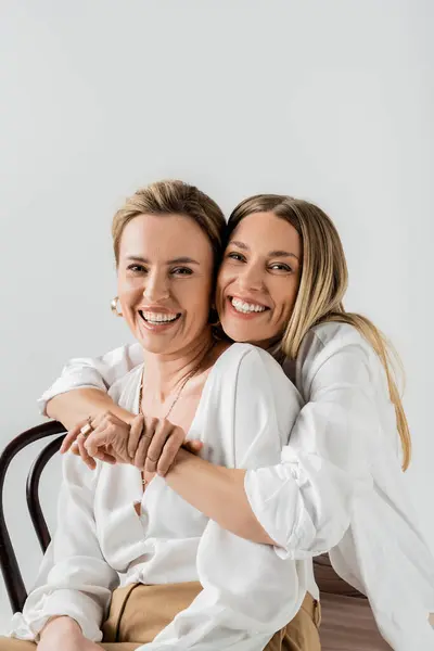 Retrato de irmãs loiras elegantes sentadas em cadeiras abraçando e sorrindo, estilo e moda — Fotografia de Stock