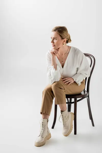 Blonde Frau in pastellfarbener Kleidung sitzt auf Stuhl und schaut weg, Hand an Kinn, Stil und Mode — Stockfoto