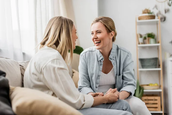 Two lovely sisters in casual wear sitting on sofa smiling and holding hands, family bonding — Stock Photo
