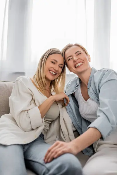 Fechar duas irmãs sentadas no sofá rindo com os olhos fechados, mão no joelho, ligação familiar — Fotografia de Stock