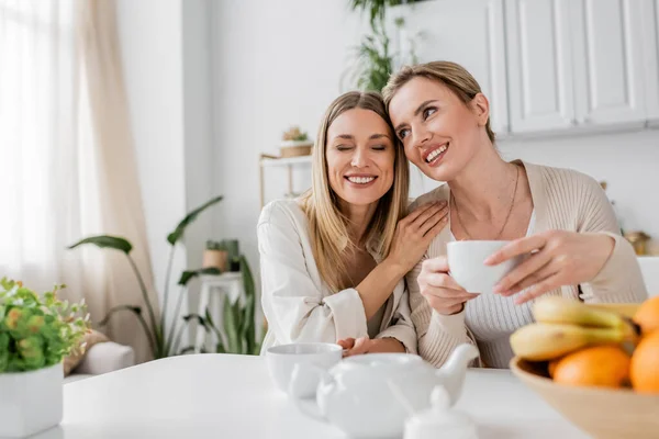 Lovely attractive sisters sitting in kitchen and smiling, hand on shoulder, family bonding — Stock Photo