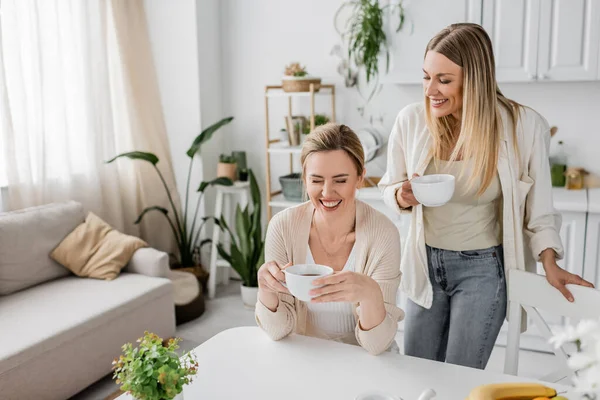 Duas irmãs bonitas bebendo chá e sorrindo no cenário de móveis de cozinha, ligação familiar — Fotografia de Stock
