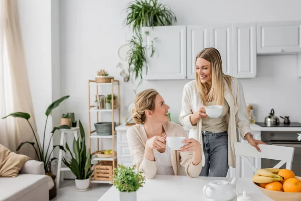 Beautiful lovely sisters smiling and looking at each other on kitchen backdrop, family bonding — Stock Photo