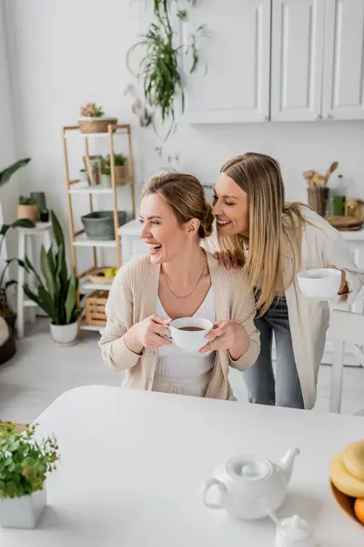 Close up cheerful sisters in casual attire smiling and looking away with kitchen backdrop, bonding — Stock Photo