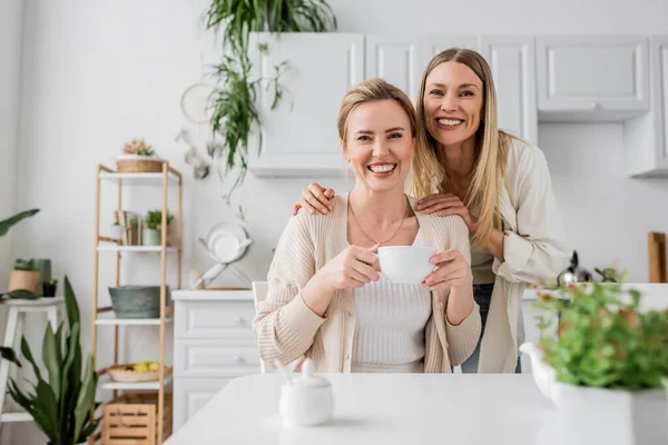 Duas irmãs loiras bebendo chá e sorrindo no fundo da cozinha com plantas, ligação familiar — Fotografia de Stock