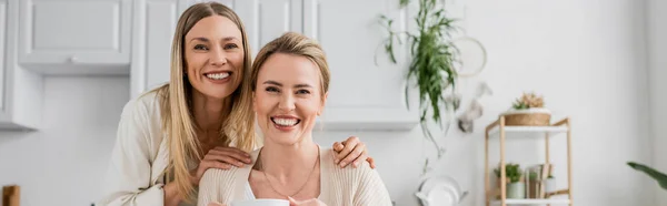 Blonde sisters smiling and looking at camera on kitchen backdrop with plants, family bonding, banner — Stock Photo