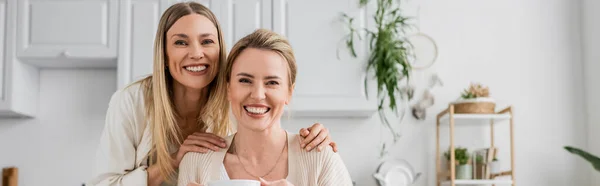 Soeurs blondes souriantes et regardant la caméra sur fond de cuisine avec des plantes, liens familiaux, bannière — Photo de stock