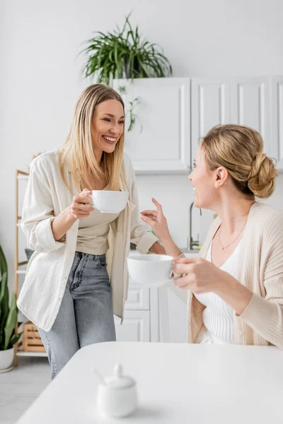 Two attractive sisters smiling and looking at each other on kitchen backdrop with plants, bonding — Stock Photo