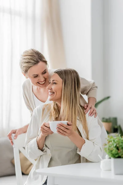 Close up two blonde sisters drinking tea and smiling on kitchen backdrop, hug from behind, bonding — Stock Photo