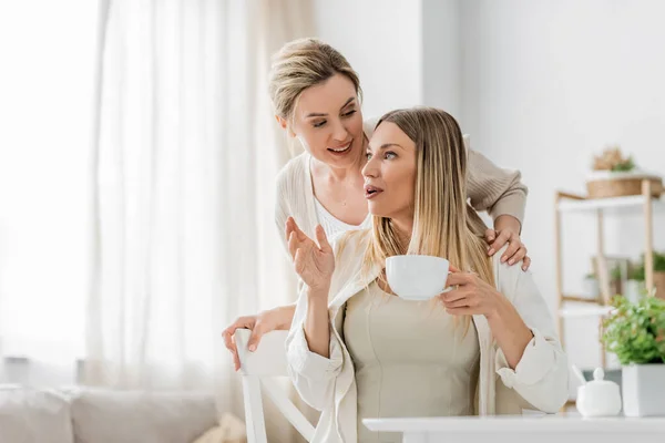 Close up blonde attractive sisters talking and smiling on kitchen backdrop, hug from behind, bonding — Stock Photo
