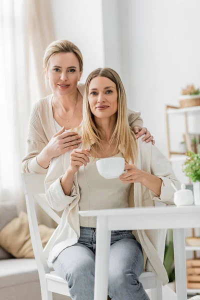 Two sisters looking at camera smiling and posing on kitchen backdrop with plants, family bonding — Stock Photo