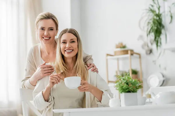 Deux jolies sœurs gaies regardant la caméra souriant et posant sur fond de cuisine, liens de famille — Photo de stock