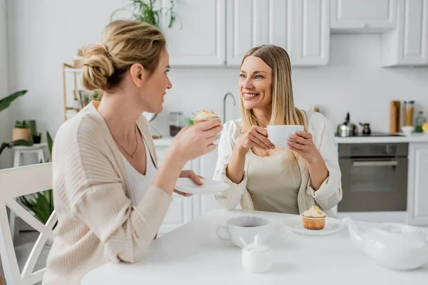 Good looking sisters drinking tea and eating cupcakes on kitchen furniture backdrop, family bonding — Stock Photo