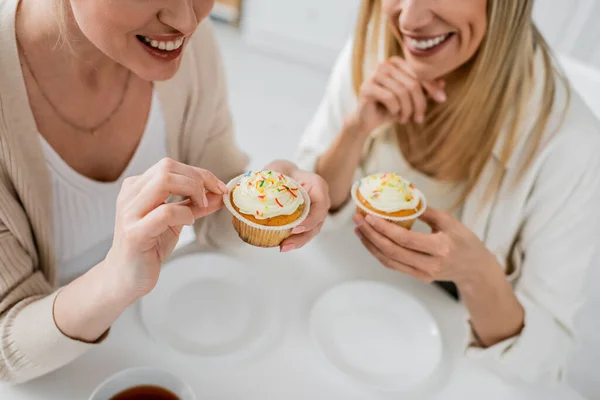 Cropped view of good looking sisters in trendy wear tasting and enjoying cupcakes, family bonding — Stock Photo