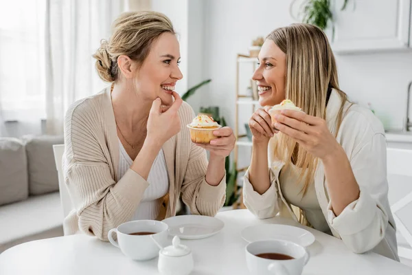 Deux jolies sœurs en tenue pastel décontractée dégustant des cupcakes et du thé sur fond de cuisine, collant — Photo de stock