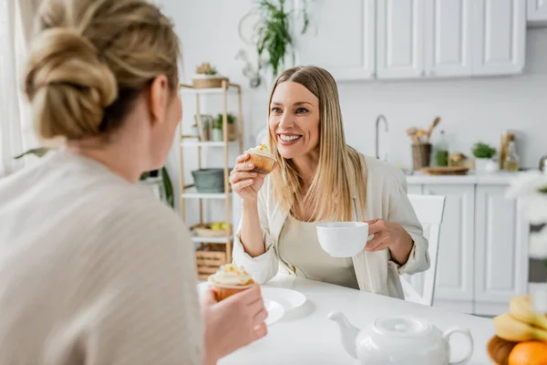 Pretty blonde sisters in white jackets enjoying tea and cupcakes and looking at each other, bonding — Stock Photo