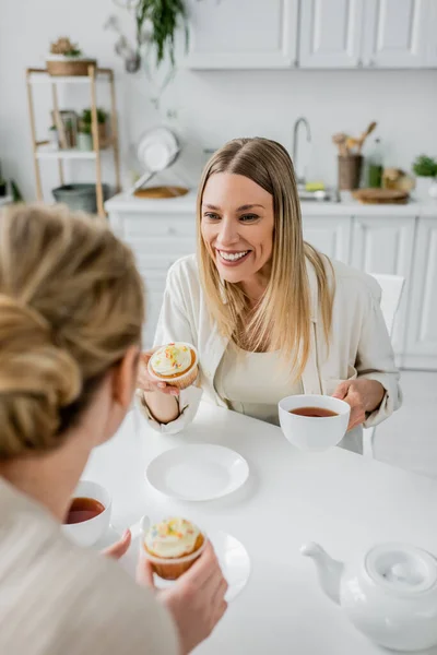 Vista cortada de duas irmãs loiras alegres comendo cupcakes e bebendo chá, ligação familiar — Fotografia de Stock