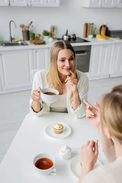 Cropped view of two blonde sisters drinking tea on kitchen backdrop with furniture, family bonding — Stock Photo