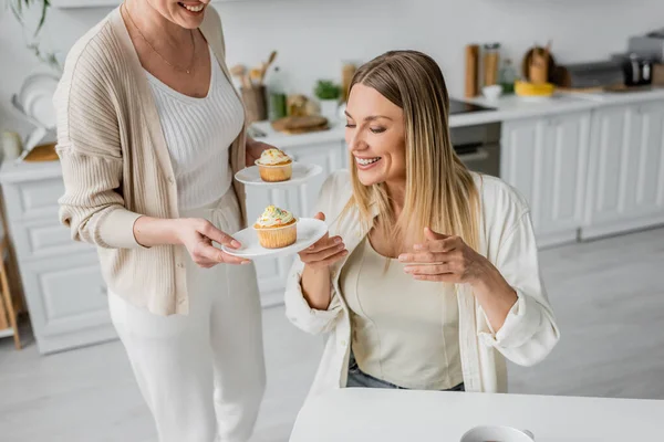 Cropped view of two sisters looking at cupcakes and smiling on kitchen backdrop, family bonding — Stock Photo