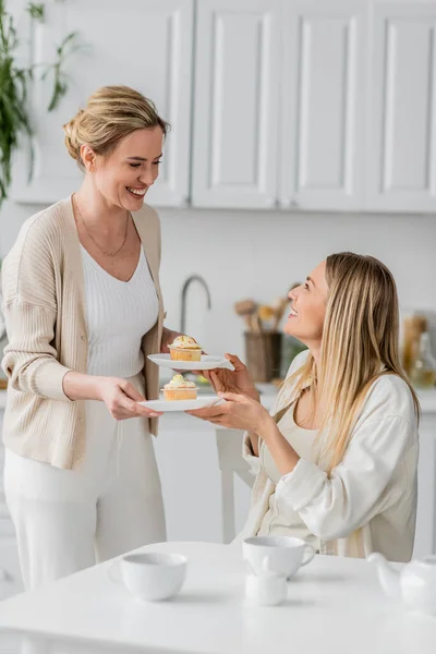 Close up cute sisters in trendy pastel cardigans holding plates with cupcakes and smiling, bonding — Stock Photo