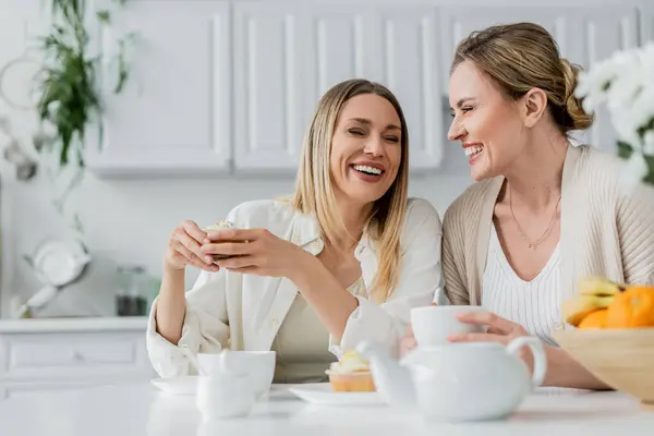 Счастливые блондинистые сестры смеются над столом и наслаждаются чаем и кексами, общением, семейными узами — стоковое фото