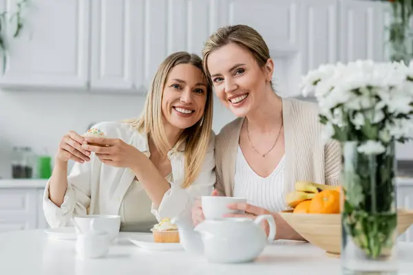 Cheerful lovely sisters in trendy attire enjoying tea and cupcakes and looking at camera, bonding — Stock Photo