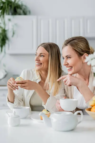 Dos hermanas rubias guapas disfrutando de cupcakes y té y mirando hacia otro lado, señalando con el dedo, uniéndose - foto de stock