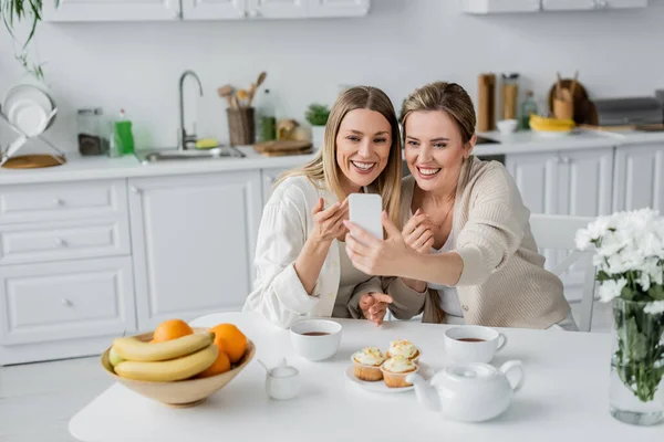 Duas irmãs em traje pastel fazendo selfie na mesa da cozinha com cupcakes e chá, ligação familiar — Fotografia de Stock