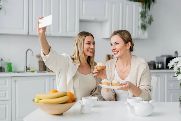 Дві модні сестри роблять селфі посміхаючись і насолоджуючись кексами на кухонному фоні, сімейне з'єднання — стокове фото