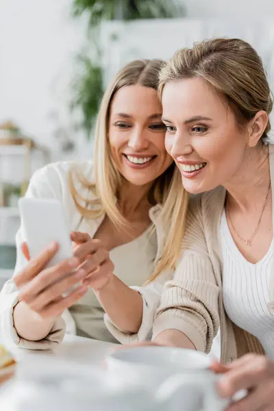 Chiudere due sorelle in abiti di bell'aspetto guardando il telefono cellulare e sorridendo, legame familiare — Foto stock