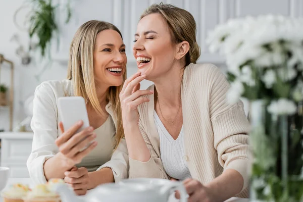 Two attractive sisters in pastel cardigans laughing and holding mobile phone, family bonding — Stock Photo