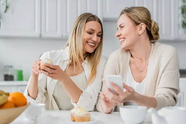 Two blonde sisters enjoying cupcakes and holding mobile phone on kitchen backdrop, family bonding — Stock Photo