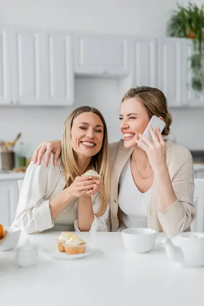 Lovely laughing sisters tasting cupcakes and holding phone on kitchen background, family bonding — Stock Photo