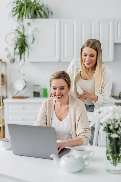 Deux soeurs blondes en tenue tendance regardant ordinateur portable sur fond de cuisine, liens familiaux — Photo de stock
