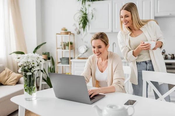 Good looking cheerful sisters in lovely attire looking at laptop and holding a tea cup, bonding — Stock Photo
