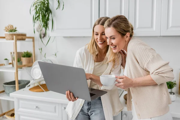 Due sorelle allegre guardando il computer portatile e sorridendo sinceramente in possesso di tazza di tè, legame familiare — Foto stock