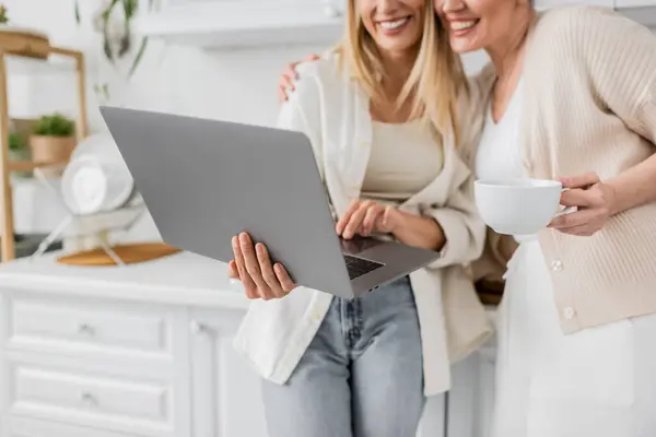 Vista recortada de dos hermanas rubias sonrientes en cardigans pastel de moda mirando el ordenador portátil, vinculación - foto de stock