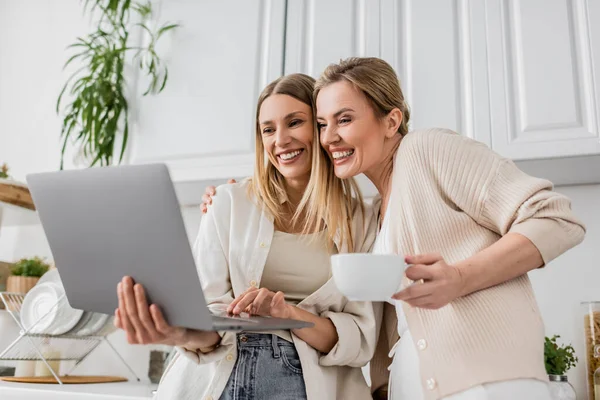 Two smiling sisters looking at laptop and hugging on kitchen backdrop, family bonding — Stock Photo